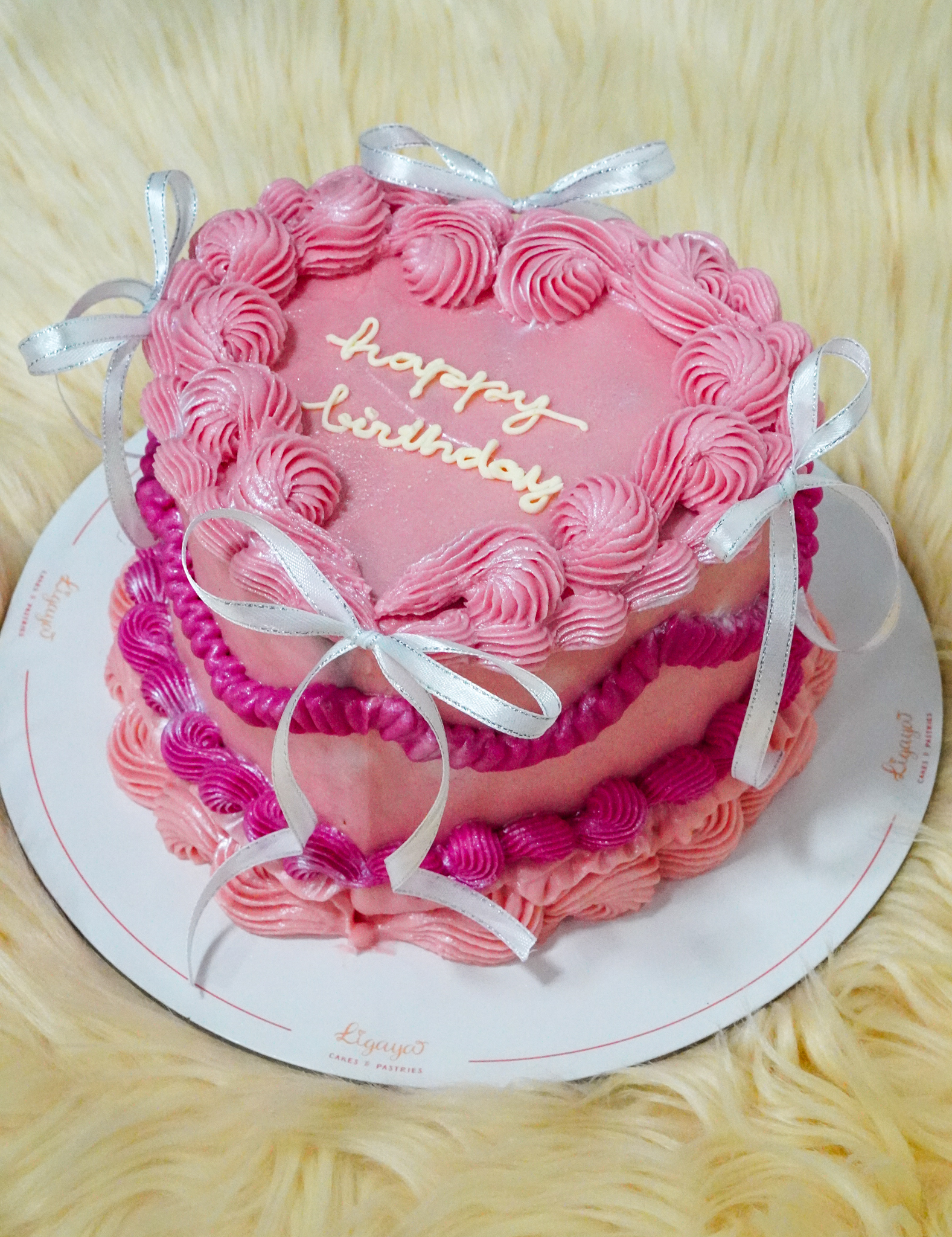 Pink heart-shaped birthday cake with two tiers, decorated with pink buttercream swirls and silver ribbon bows