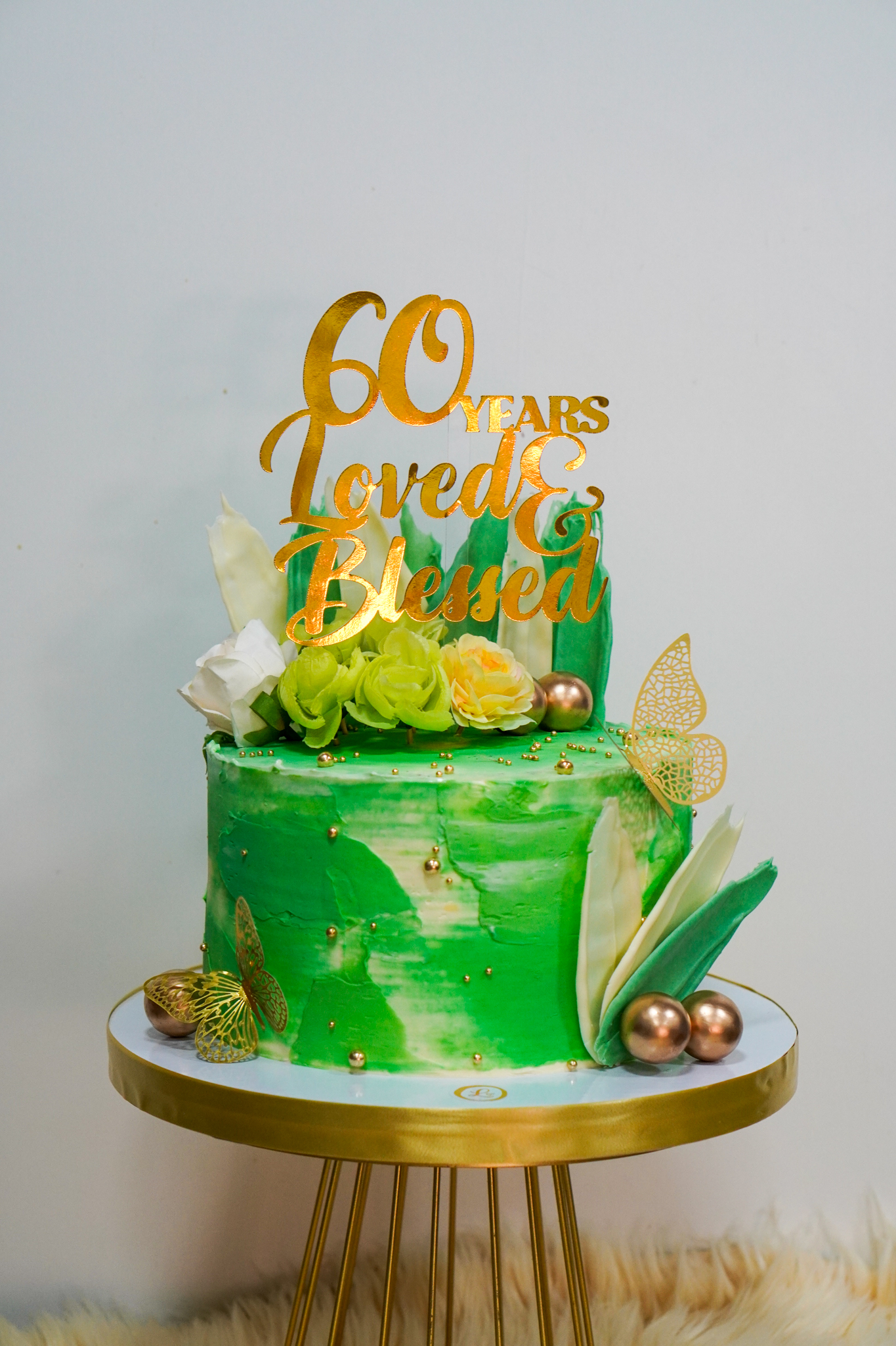Green and gold birthday cake with fresh flowers, decorative butterflies, and gold topper