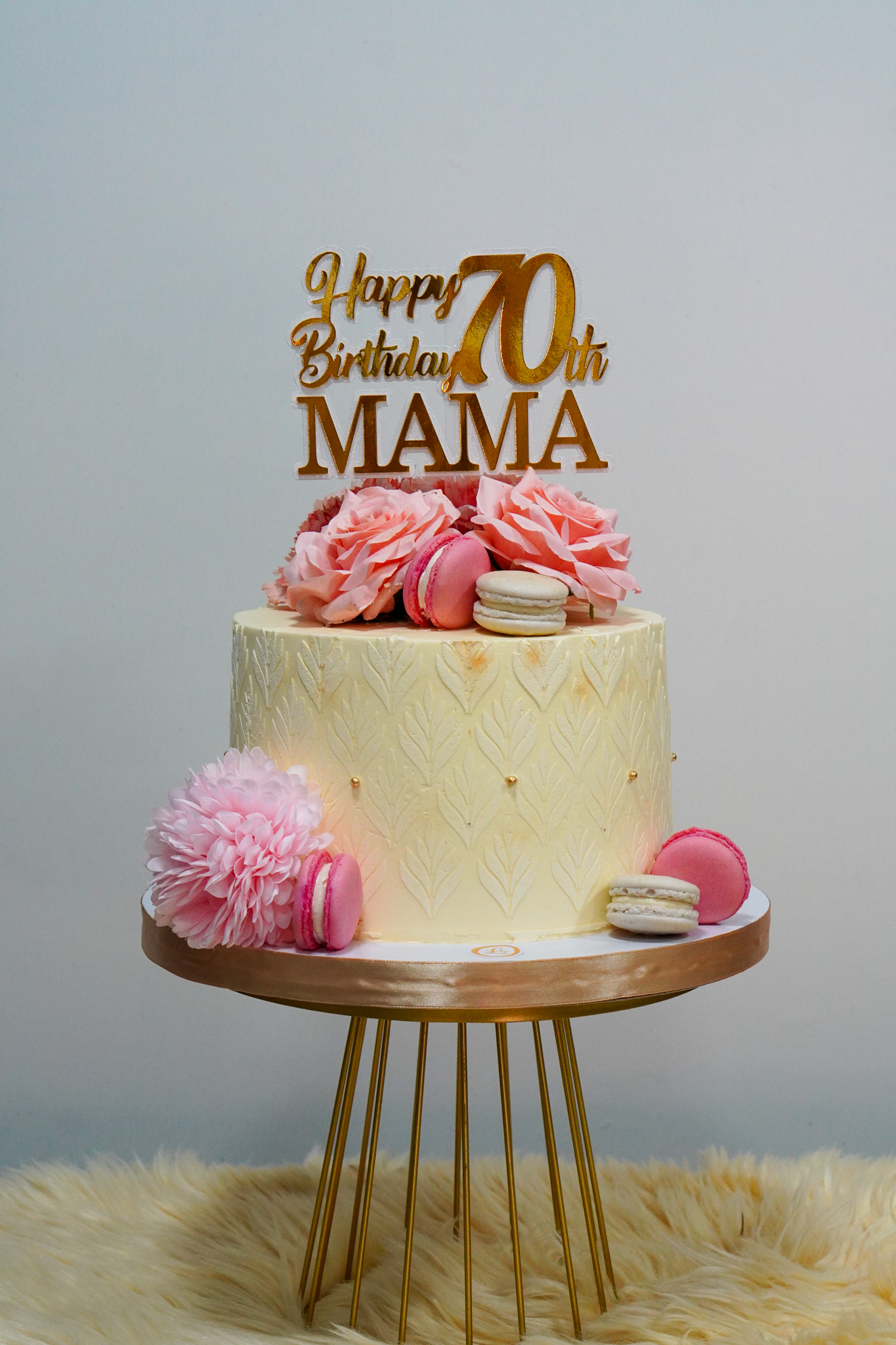 Elegant cream birthday cake with pink flowers, macarons, and gold topper for a 70th birthday