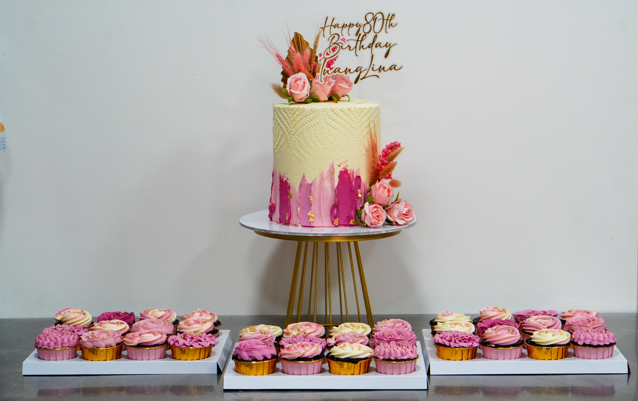 Elegant cream and pink floral birthday cake with textured design, accompanied by matching pink and cream cupcakes