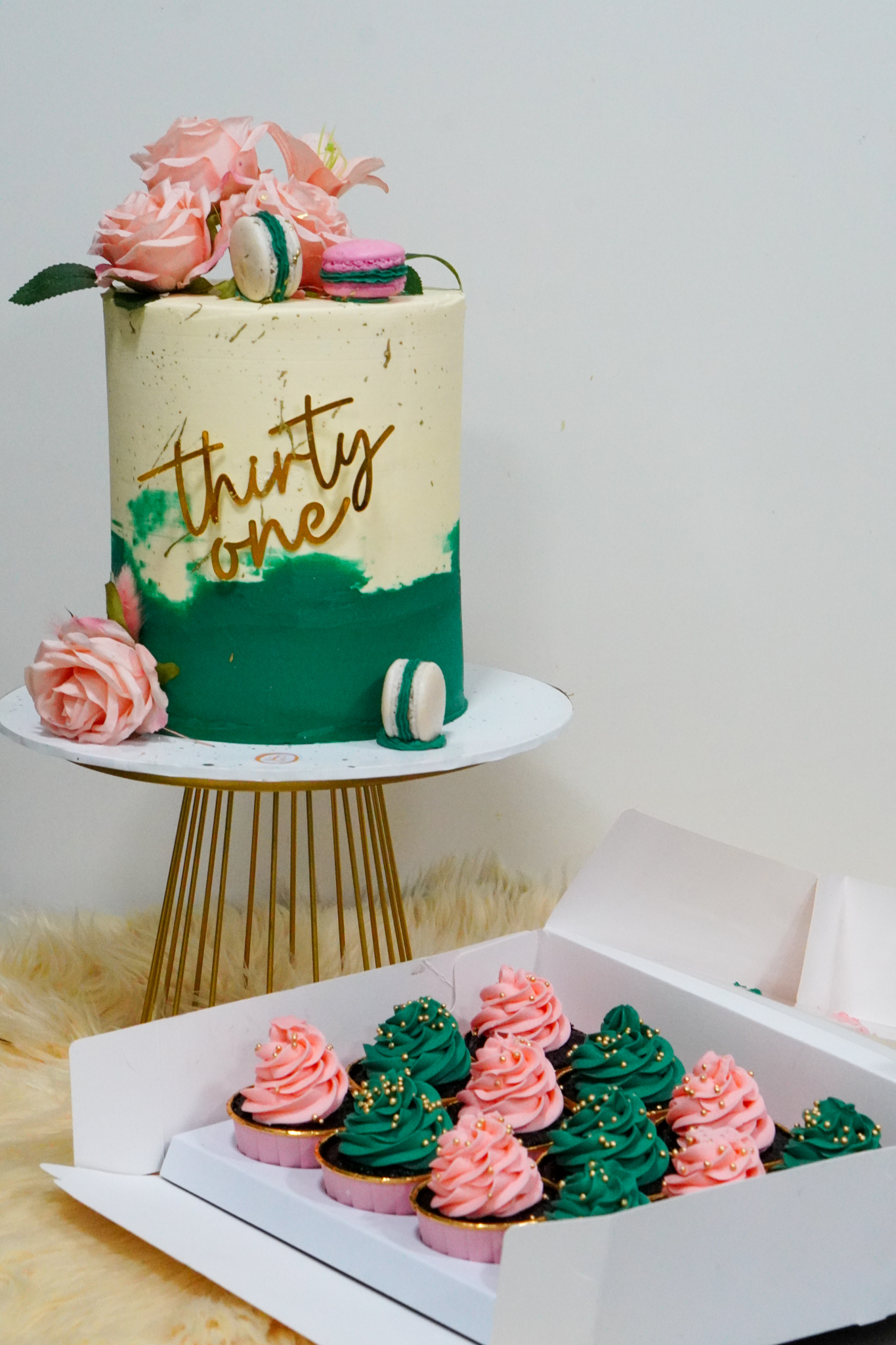 Elegant green and cream birthday cake with 'thirty one' topper, pink roses, and macarons, paired with matching pink and green frosted cupcakes