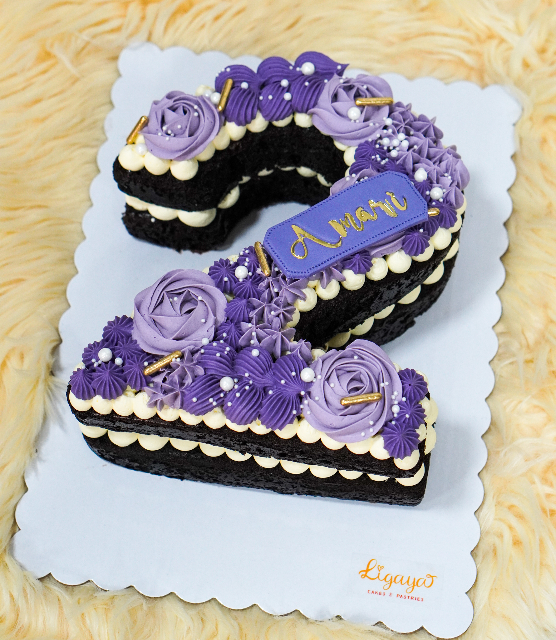 Number-shaped chocolate cake decorated with purple and white buttercream flowers, edible pearls, and gold sprinkles, with a personalized name plaque