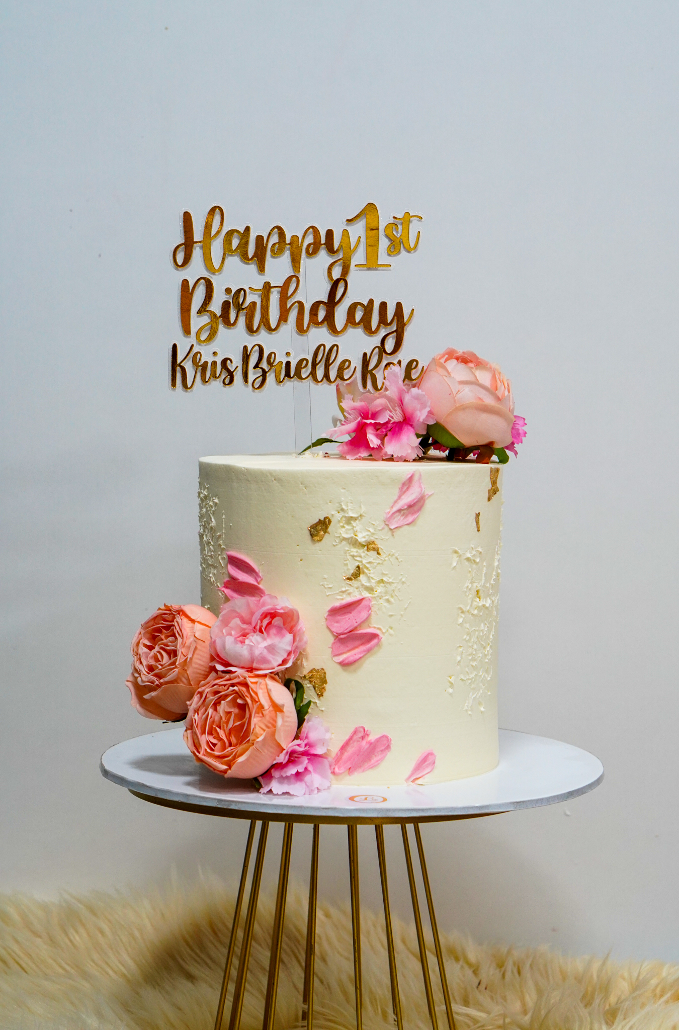 Elegant floral 1st birthday cake in ivory with pink and peach roses, pink petals, gold leaf accents, and a gold topper reading 'Happy 1st Birthday Kris Brielle Rae'