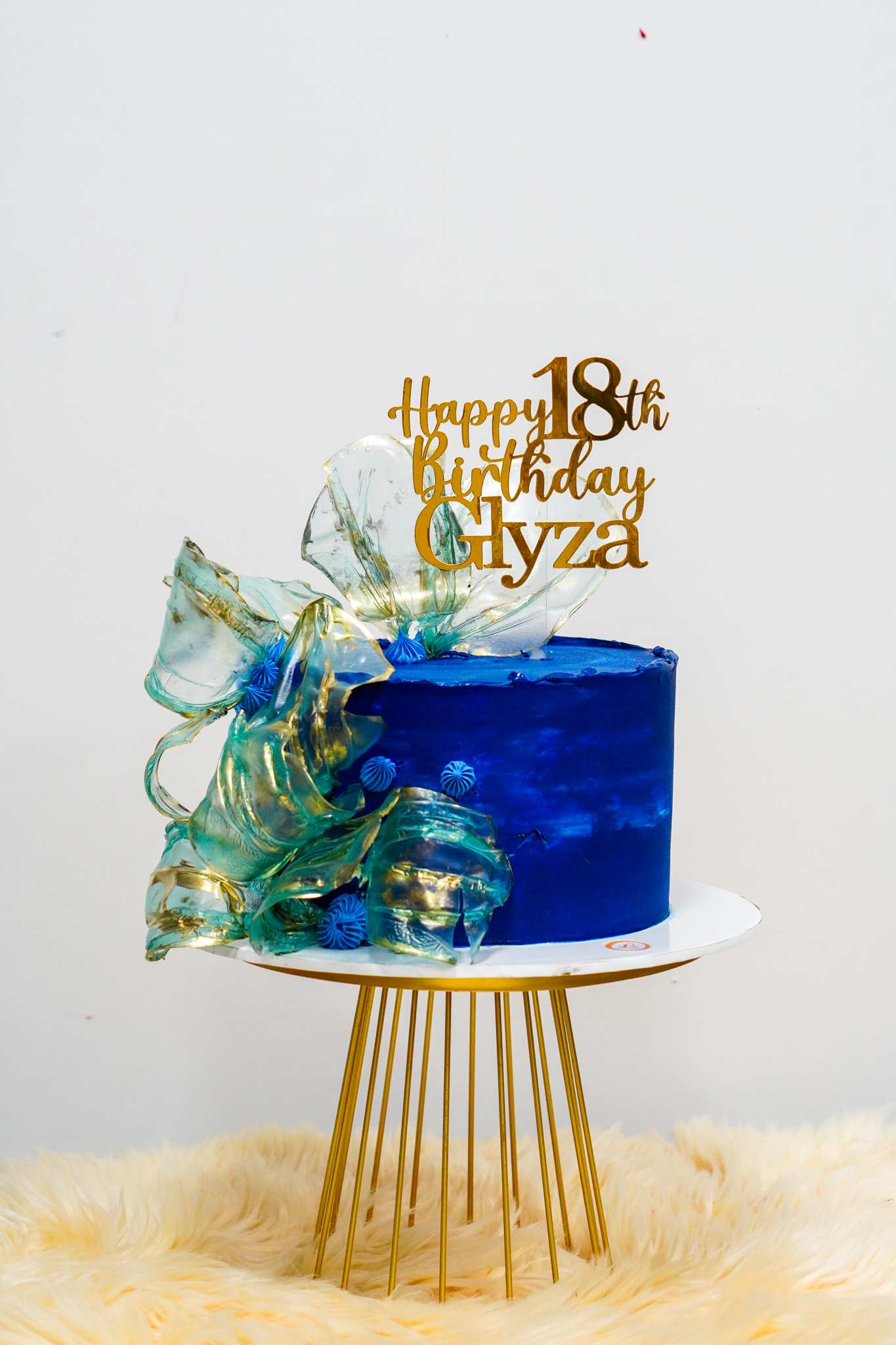 Elegant ocean-inspired 18th birthday cake in deep blue with gold accents, adorned with translucent blue sugar sails, small blue rosettes, and a gold topper reading 'Happy 18th Birthday Glyza'