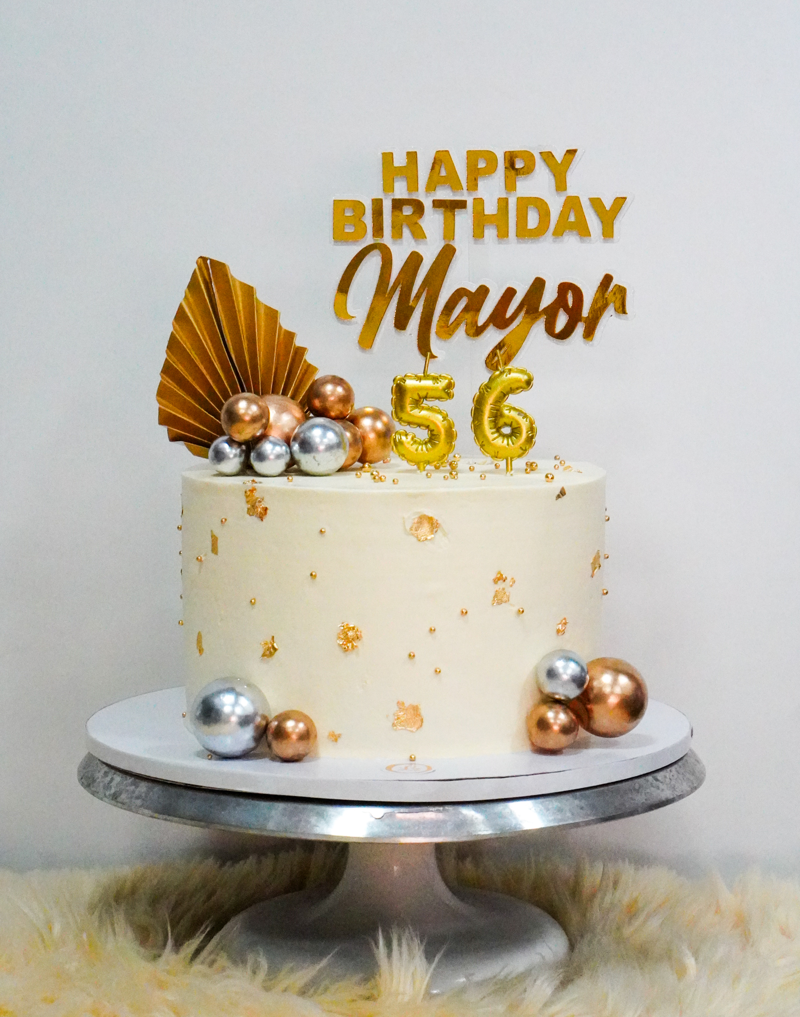 Elegant white buttercream birthday cake decorated with gold leaf accents, metallic gold, silver, and bronze edible spheres, topped with a gold topper reading 'Happy Birthday Mayor 56' and a gold fan accent