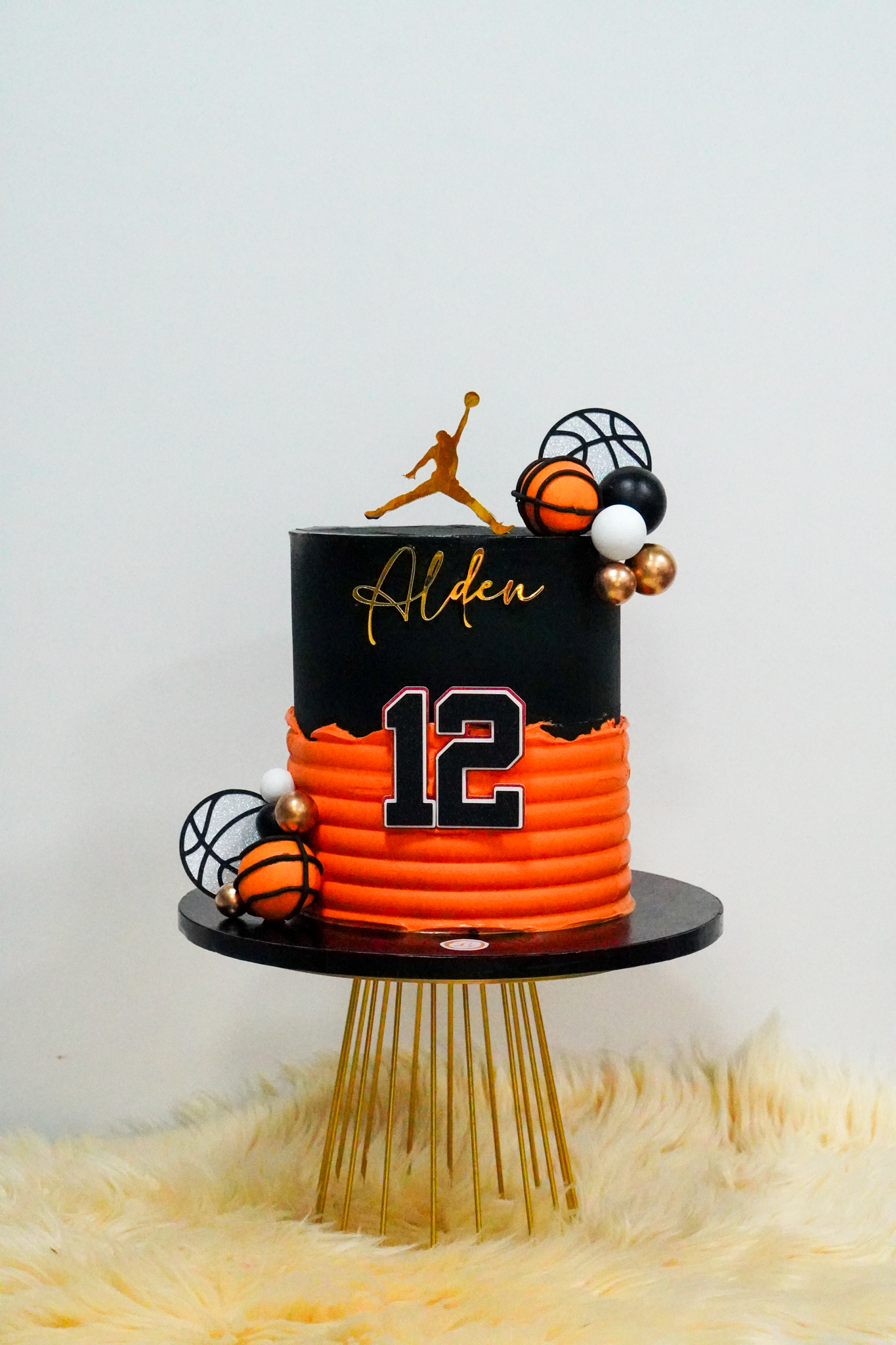Basketball-themed birthday cake with a black and orange buttercream design, topped with a gold basketball player silhouette, edible basketball decorations, metallic spheres, and the number 12 on the front with the name Alden