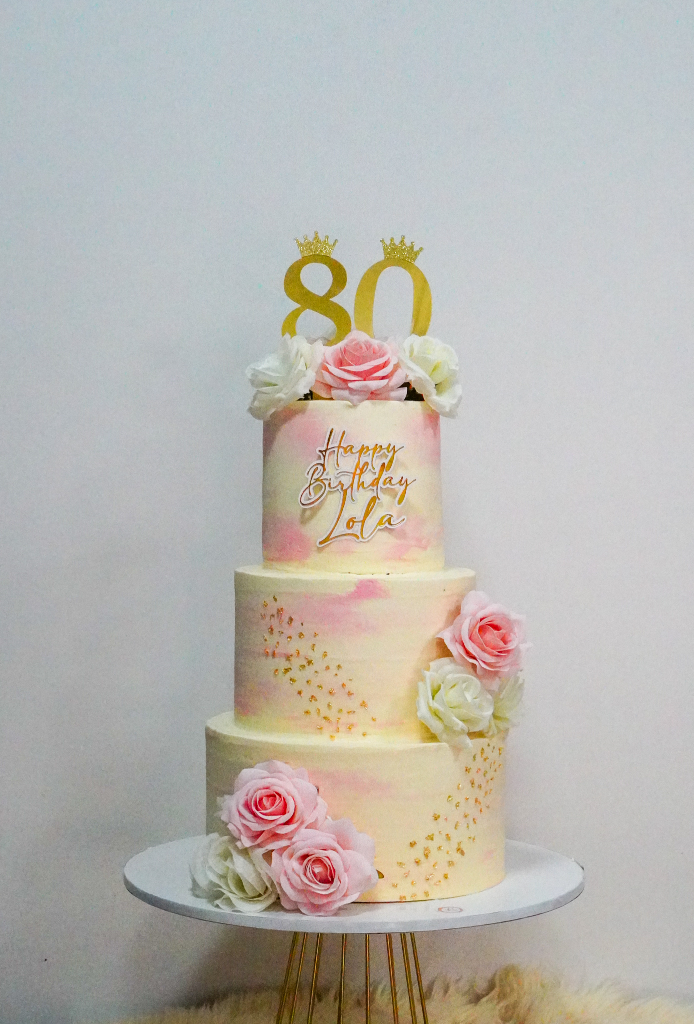 Three-tier 80th birthday cake for Lola with soft pastel pink and yellow watercolor buttercream, adorned with pink and white roses, gold crown toppers on the number 80, and delicate gold bead arrangements