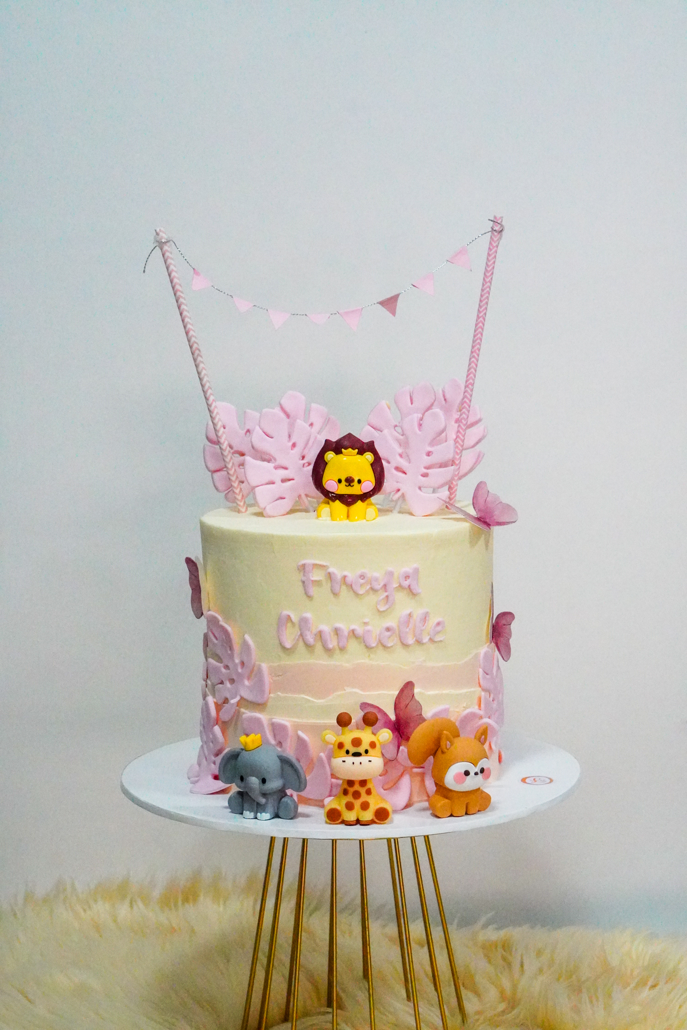 Pastel jungle-themed birthday cake with light pink and cream frosting, decorated with pink fondant leaves, edible animal toppers including a lion, elephant, giraffe, and fox, with a mini pink bunting topper, and the name 'Freya Chrielle' on the front