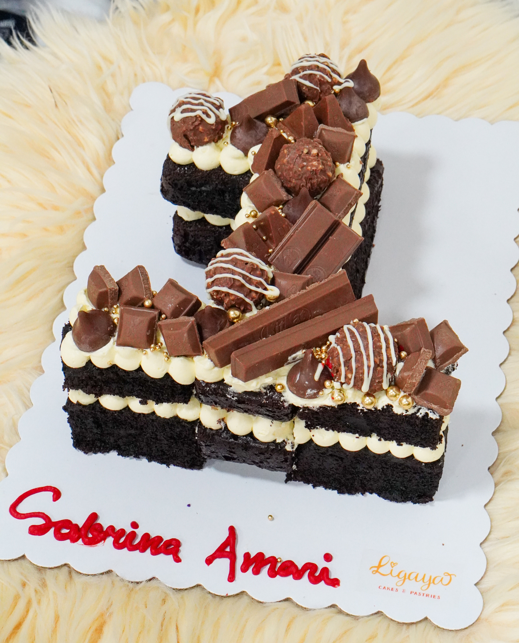 Number-shaped chocolate cake topped with assorted chocolates including KitKat, Ferrero Rocher, and chocolate-dipped strawberries, with gold pearl sprinkles
