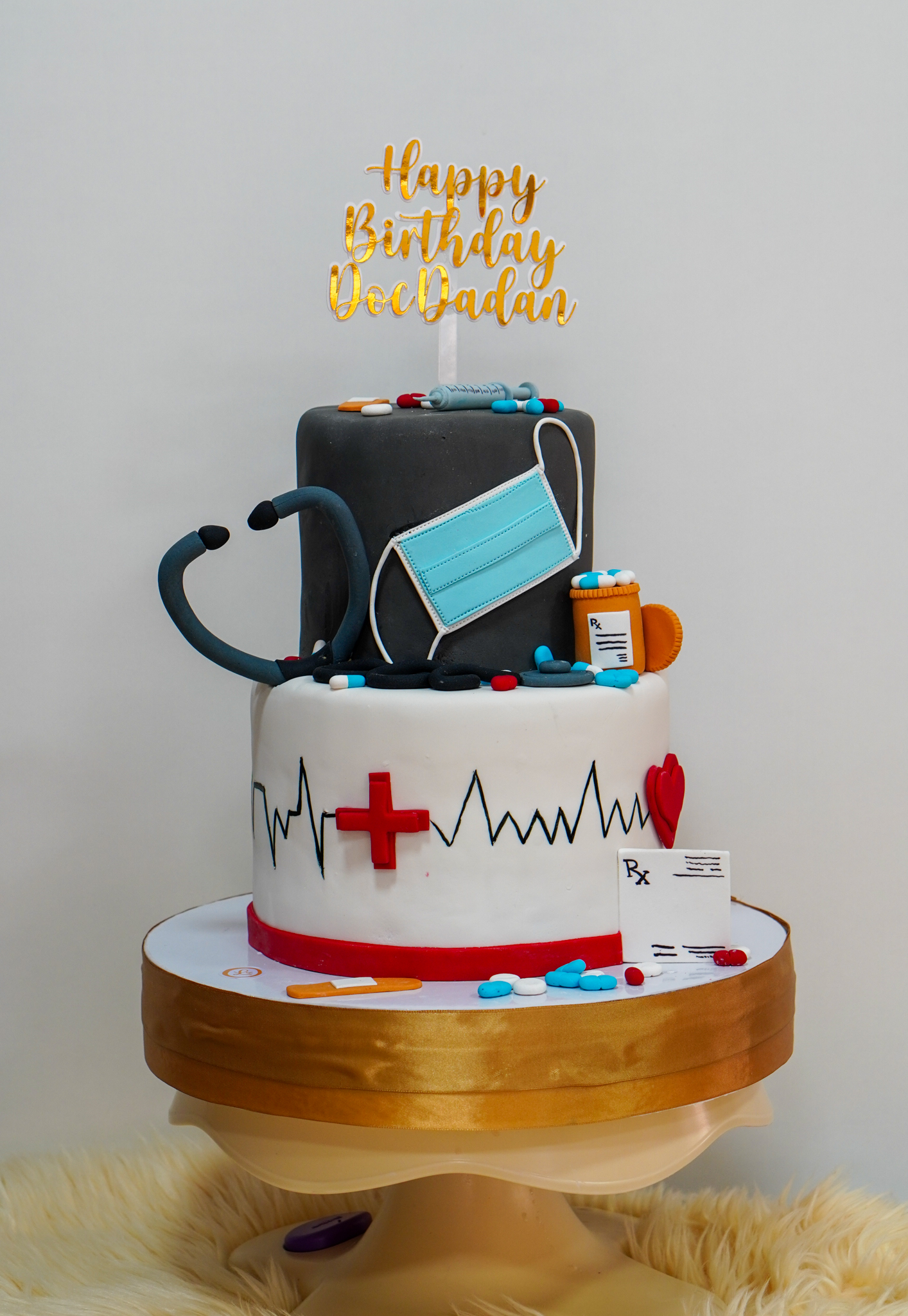 Two-tier fondant birthday cake with medical theme, featuring stethoscope, face mask, pill bottles, pills, syringe, heartbeat line, red cross, and a gold 'Happy Birthday Doc Dadan' topper
