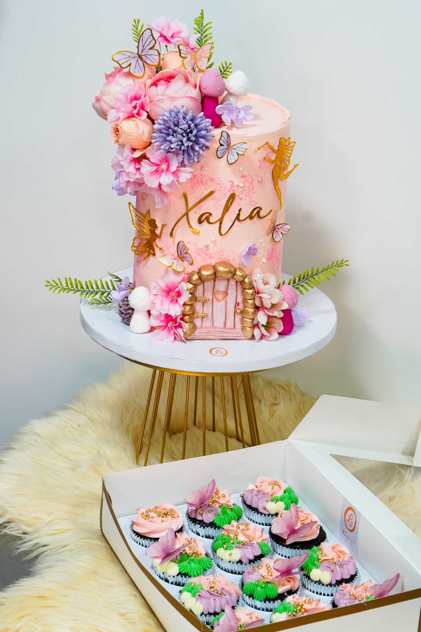 Pink buttercream fairy garden cake with gold name 'Xalia', floral decorations, butterflies, golden door design, and matching pastel cupcakes