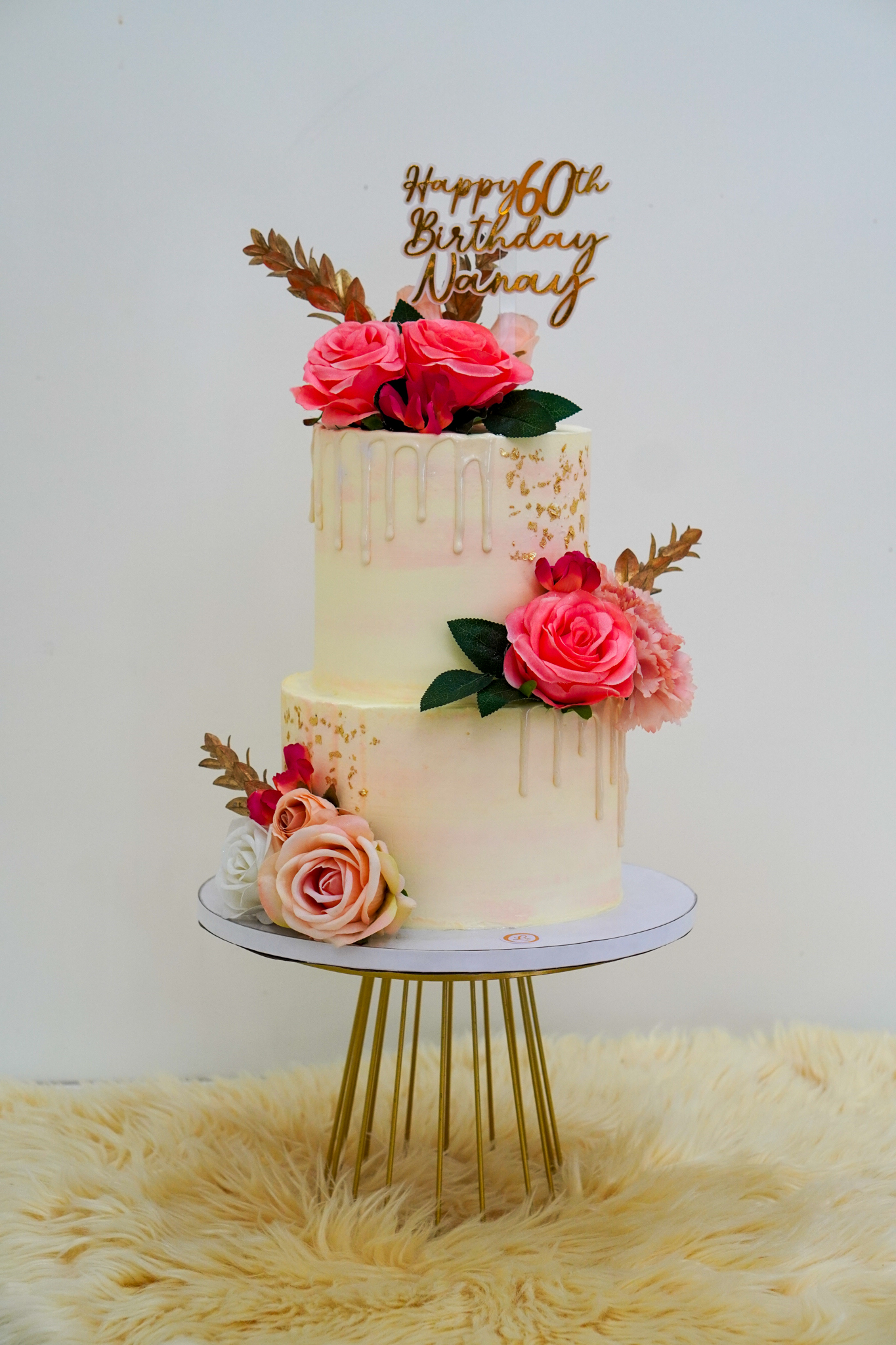 Two-tier buttercream cake in pastel pink and cream with gold drip design, decorated with pink and peach roses, gold leaves, and a gold 'Happy 60th Birthday Nanay' topper