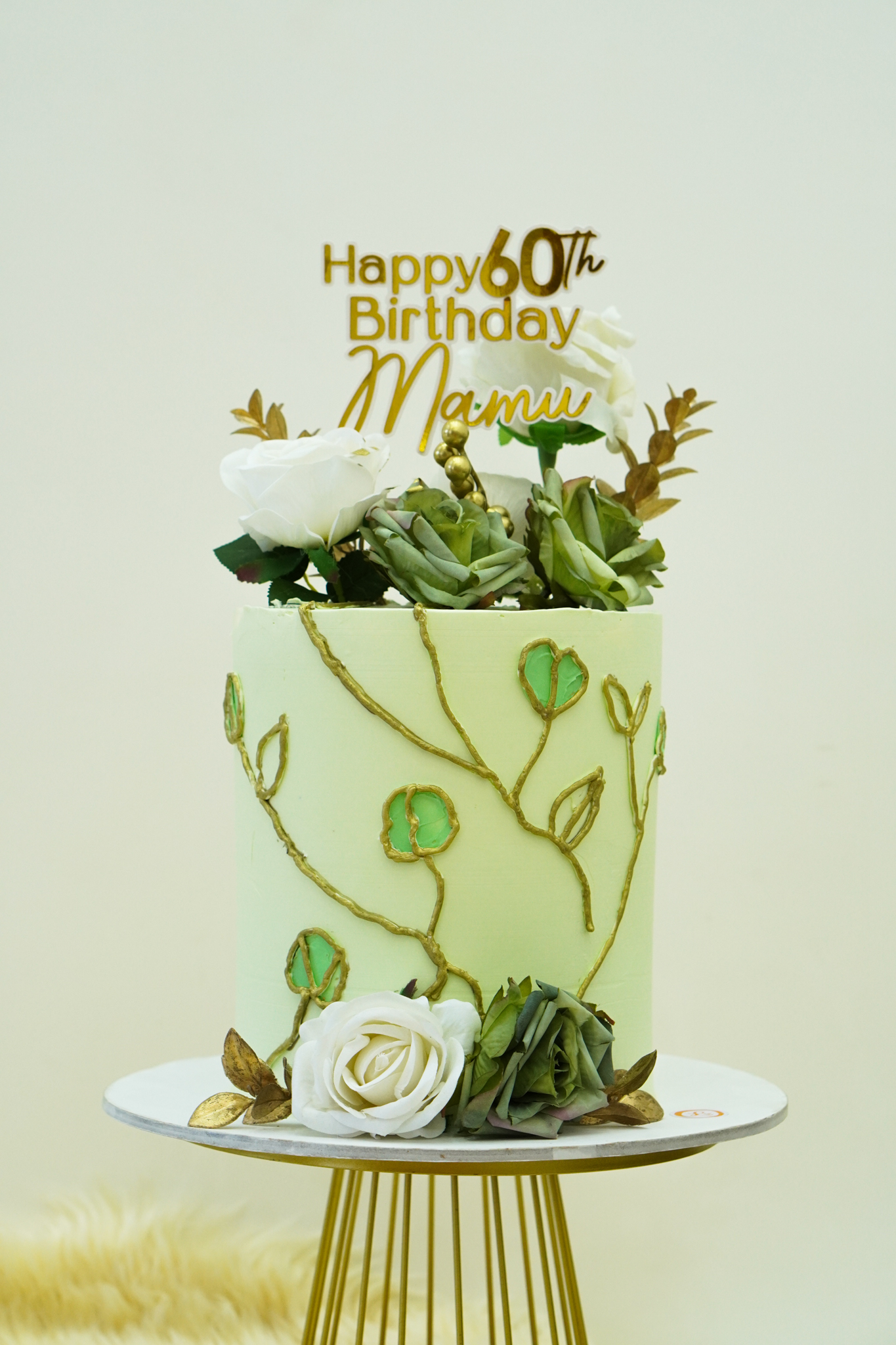 Light green buttercream birthday cake with gold vine and leaf design, topped with white and green flowers, gold leaves, and a gold 'Happy 60th Birthday Mamu' topper