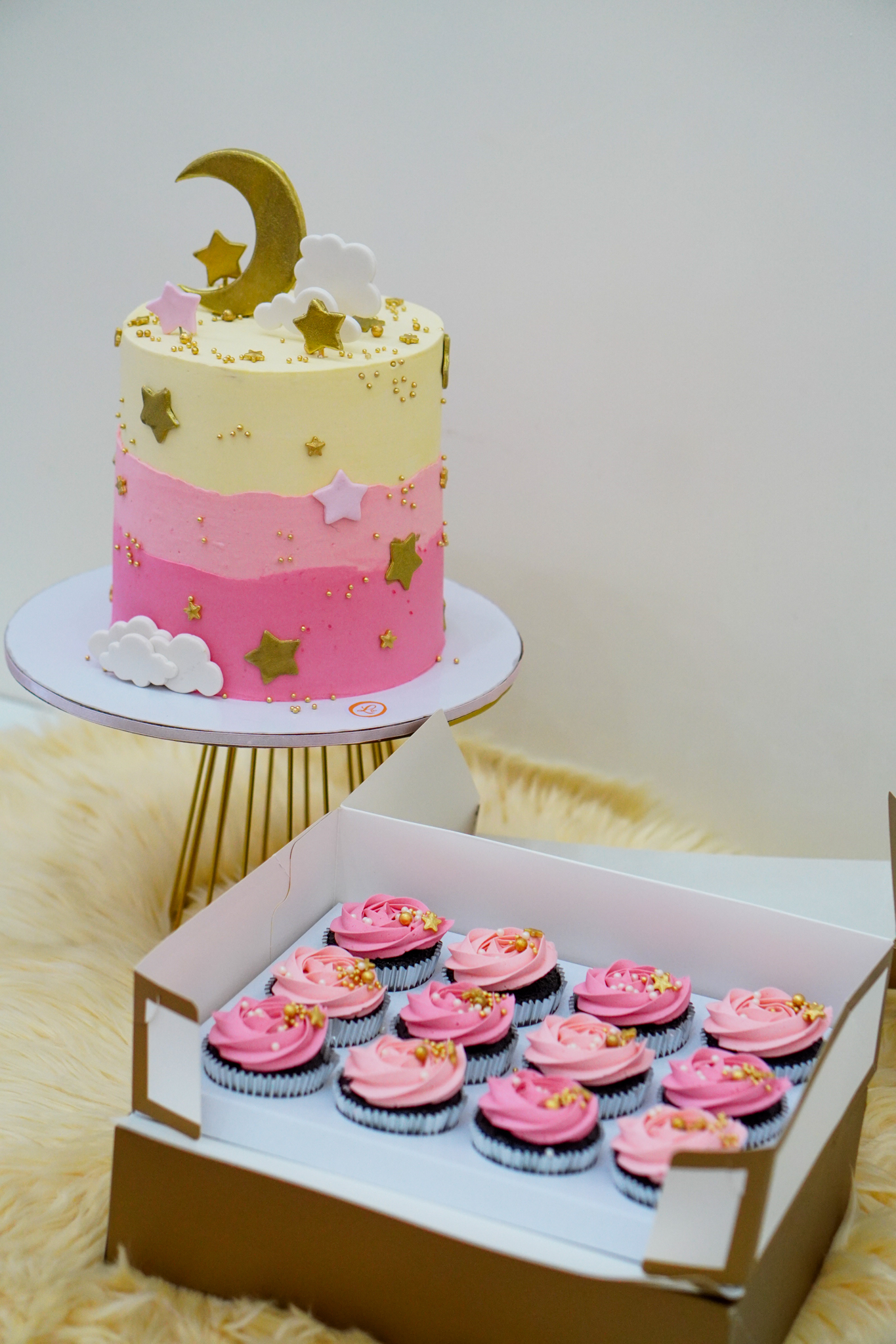 Two-tone pink and cream buttercream cake with gold crescent moon, stars, clouds, and matching pink frosted cupcakes