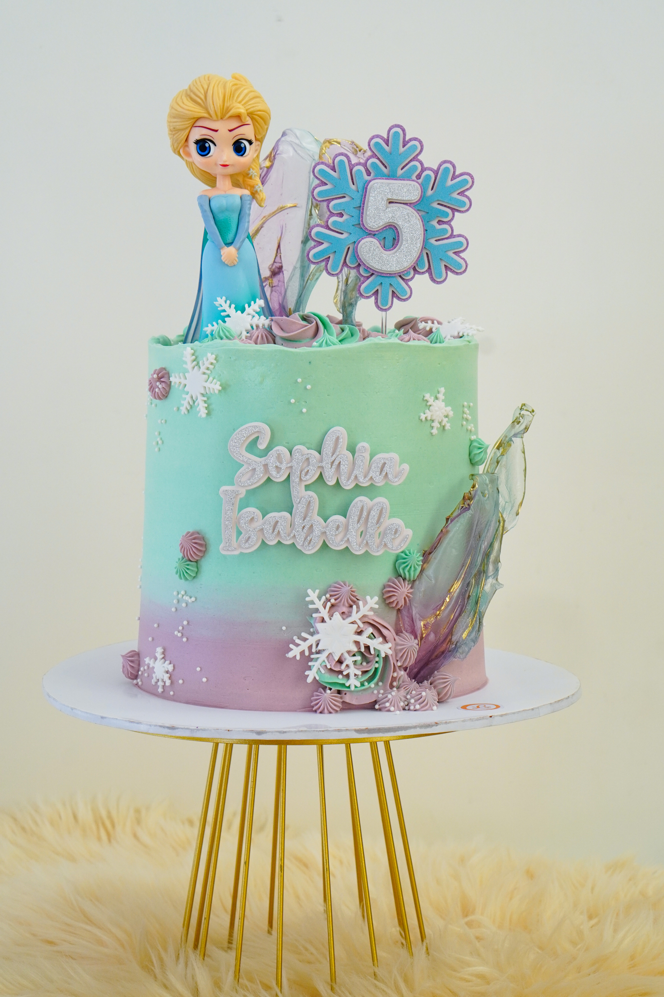 Frozen themed birthday cake in mint green and lavender ombré with Elsa figurine, snowflakes, ice shard decorations, and text 'Sophia Isabelle' with a large snowflake number 5 topper