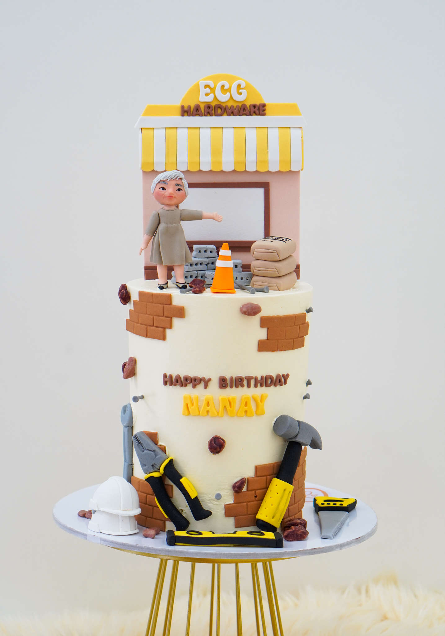 Hardware store themed buttercream birthday cake with 'ECG Hardware' storefront topper, elderly woman figurine, construction tools, bricks, and text 'Happy Birthday Nanay'
