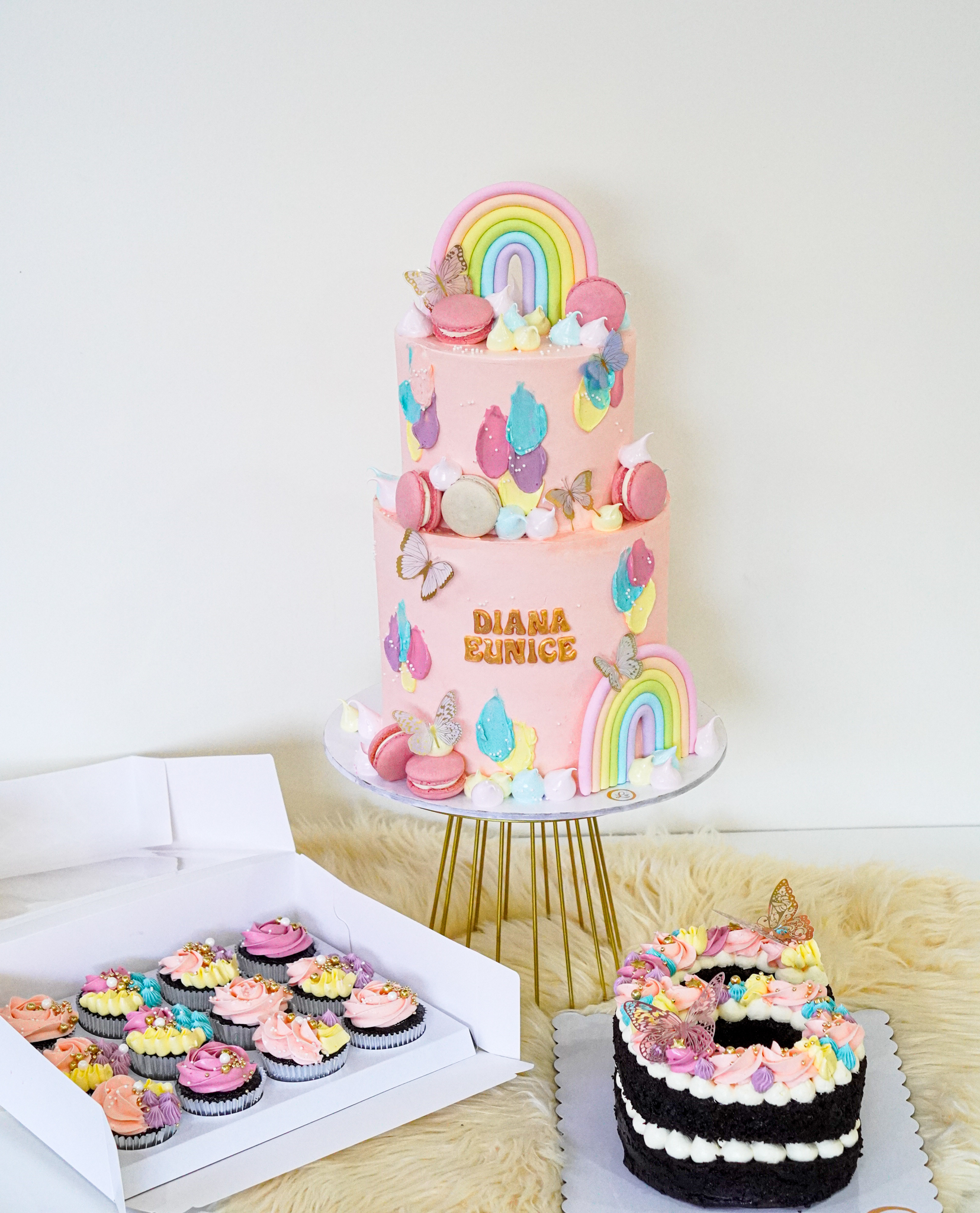 Two-tier pink buttercream birthday cake with rainbow toppers, macarons, butterflies, and colorful icing swirls, text 'Diana Eunice', accompanied by matching cupcakes and a number-shaped cake