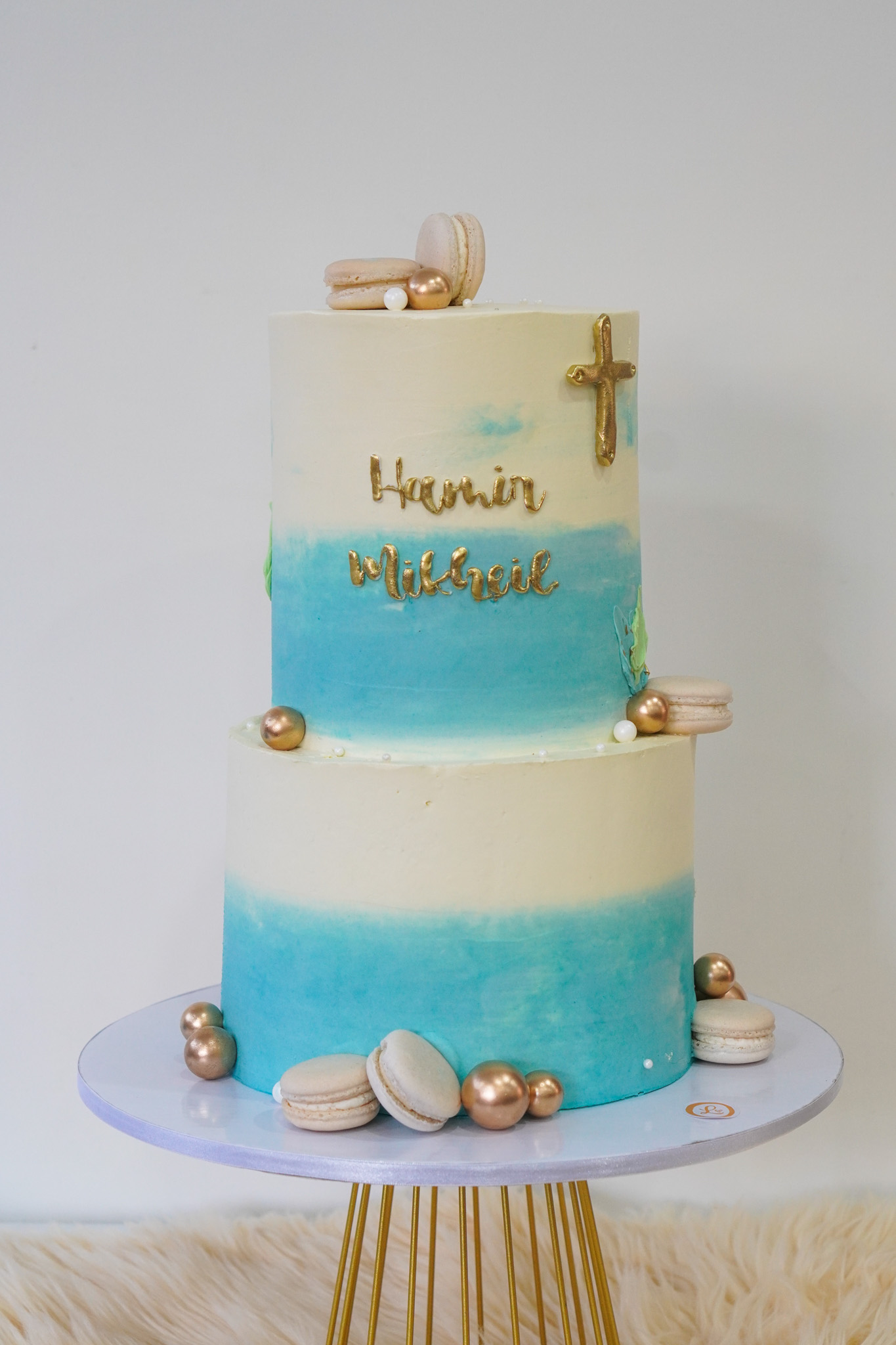 Two-tier blue ombré buttercream cake with gold cross, gold text 'Hamir Milbreil', decorated with macarons and gold pearls